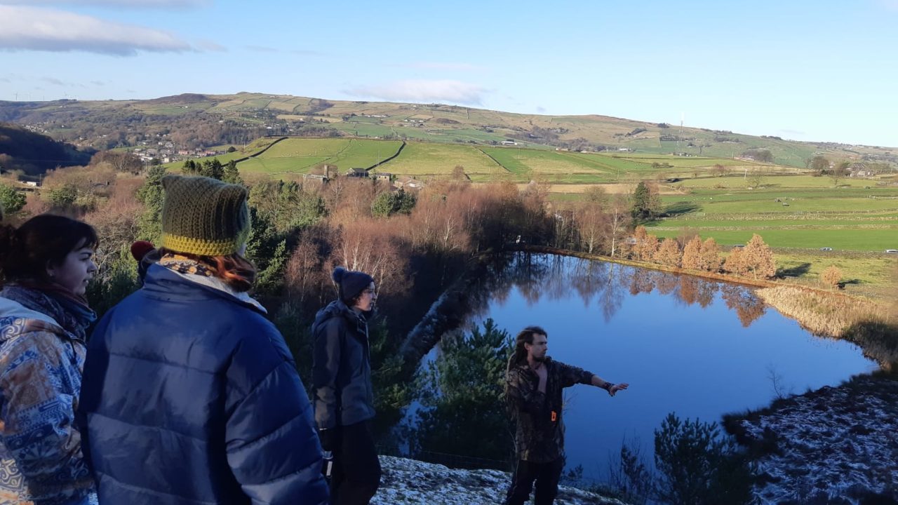 This is a landscape scene with people in it. There is a reservoir surrounded by hills. The students have their backs to the camera and Mike, a white man with long blond dreadlocks, is talking about the landscape and the improvements that have been made to Incredible Farm.