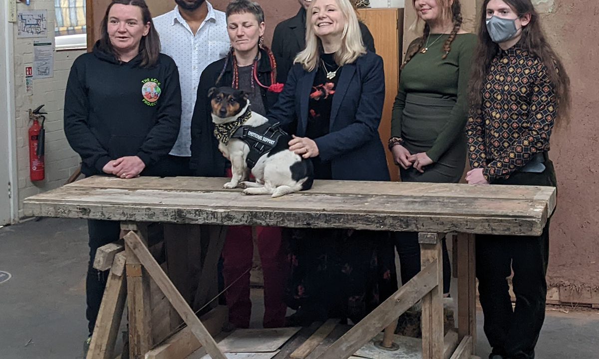 A picture (from L-R) of Anni, Alvin, Laura, the Tracy Brabin the Mayor of West Yorkshire, Cllr Scott Patient, Lola, and Evan in the workshop space with strawbale insulation blocks in the background. Reggie, a Jack Russell terrier, is on the workbench and Tracy is stroking him.