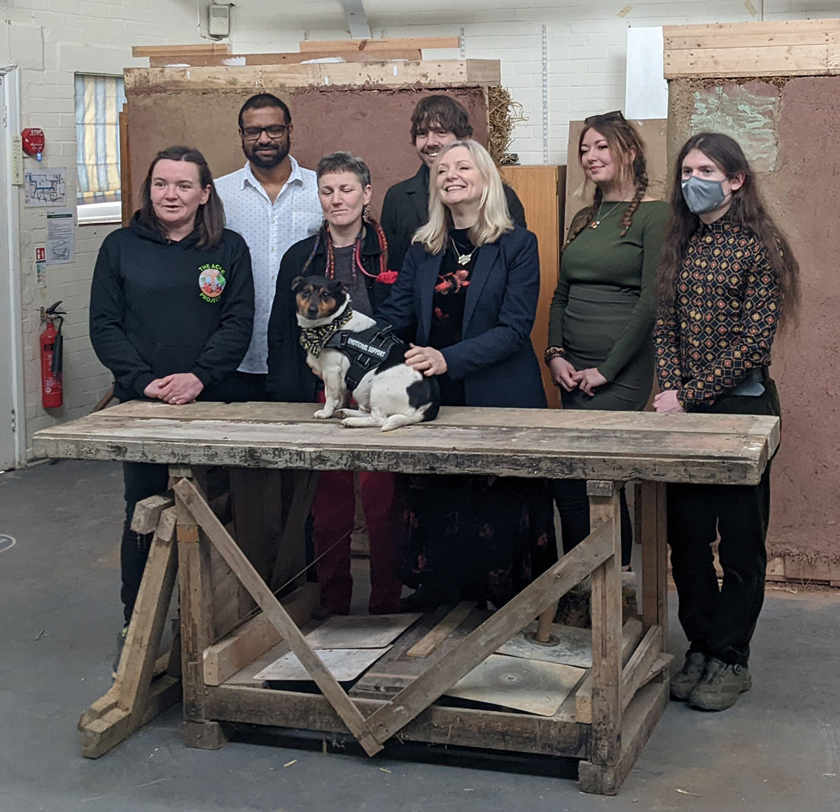 A picture (from L-R) of Anni, Alvin, Laura, the Tracy Brabin the Mayor of West Yorkshire, Cllr Scott Patient, Lola, and Evan in the workshop space with strawbale insulation blocks in the background. Reggie, a Jack Russell terrier, is on the workbench and Tracy is stroking him.