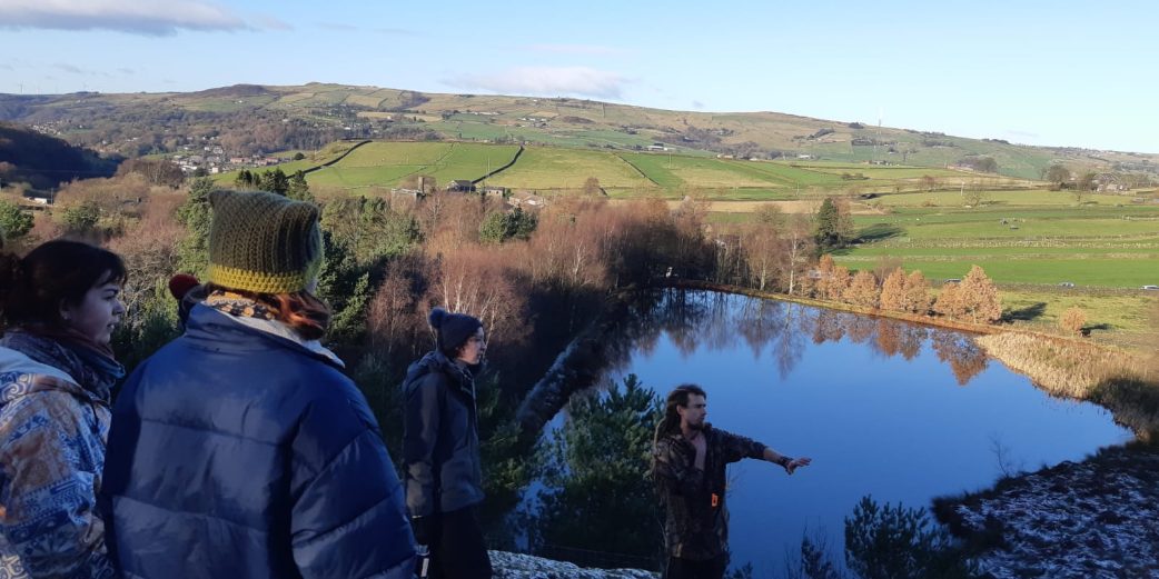 This is a landscape scene with people in it. There is a reservoir surrounded by hills. The students have their backs to the camera and Mike, a white man with long blond dreadlocks, is talking about the landscape and the improvements that have been made to Incredible Farm.